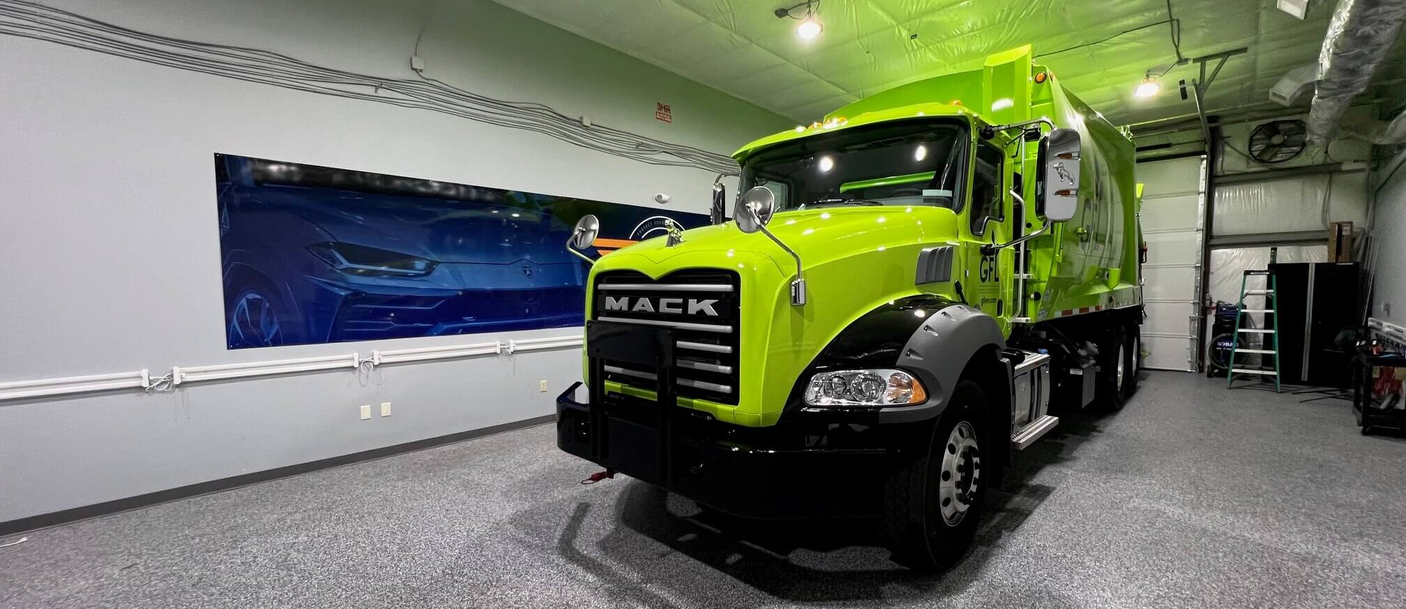 A bright green Mack garbage truck parked inside a large, well-lit garage with a high insulated ceiling and polished gray flooring. A blue mural of a car is visible on the left wall, and a ladder and equipment are seen near the back wall