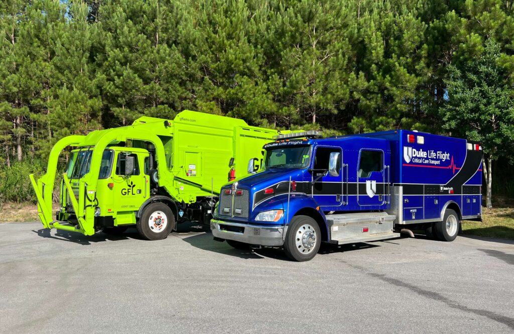 A bright green GFL Environmental garbage truck parked beside a blue Duke Life Flight critical care transport ambulance on a paved lot with a backdrop of tall pine trees