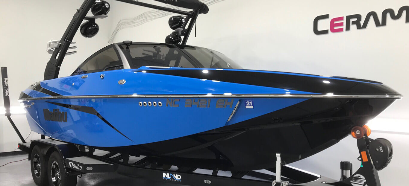 Glossy blue and black Malibu boat on a trailer inside a detailing garage with Ceramic Pro branding on the wall.