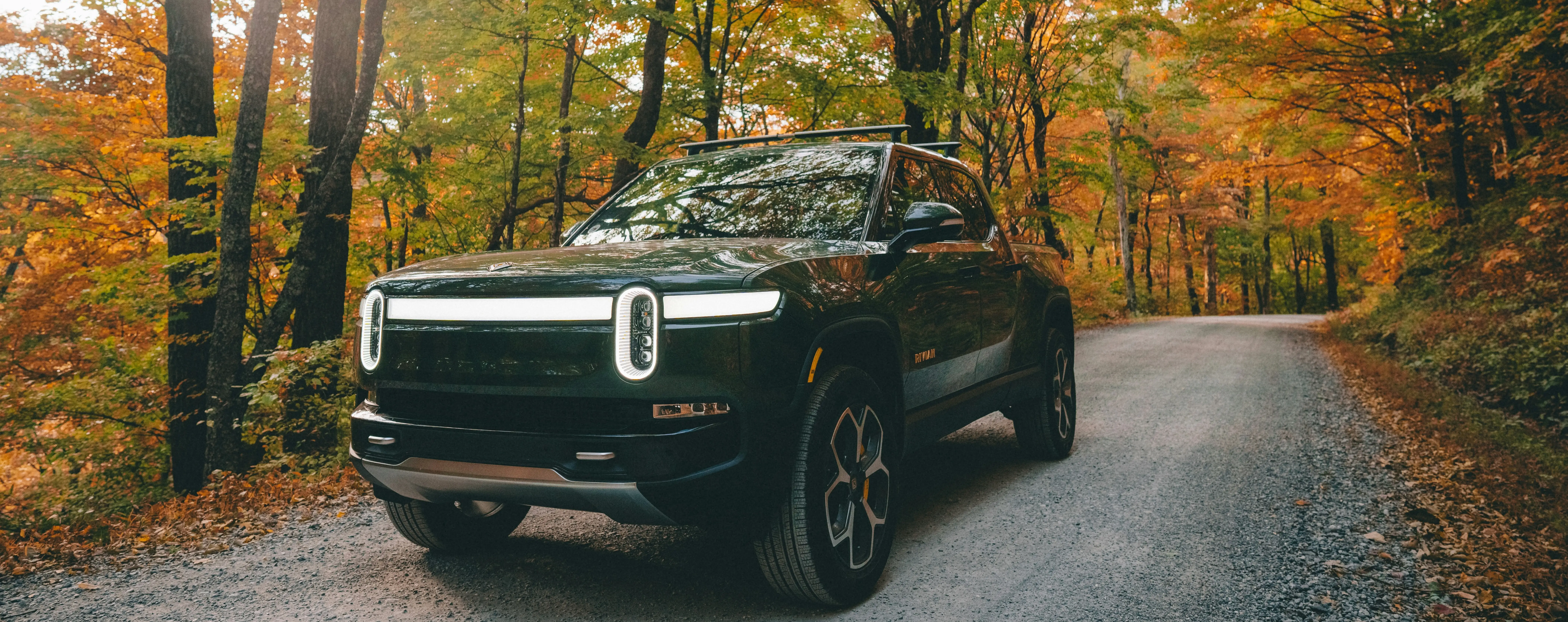 Electric pickup truck parked on a gravel road surrounded by colorful autumn forest foliage.