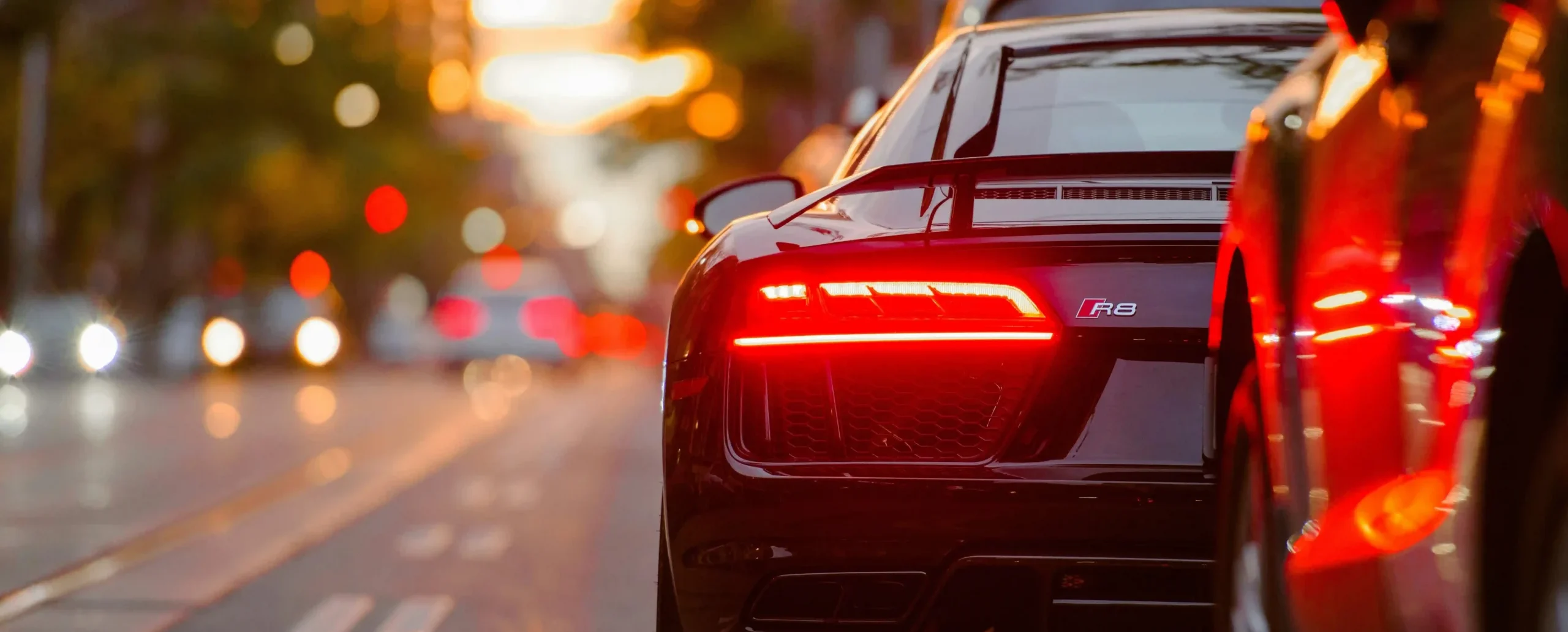 Rear view of a black Audi R8 sports car driving through a city street at sunset with glowing taillights and urban traffic bokeh.